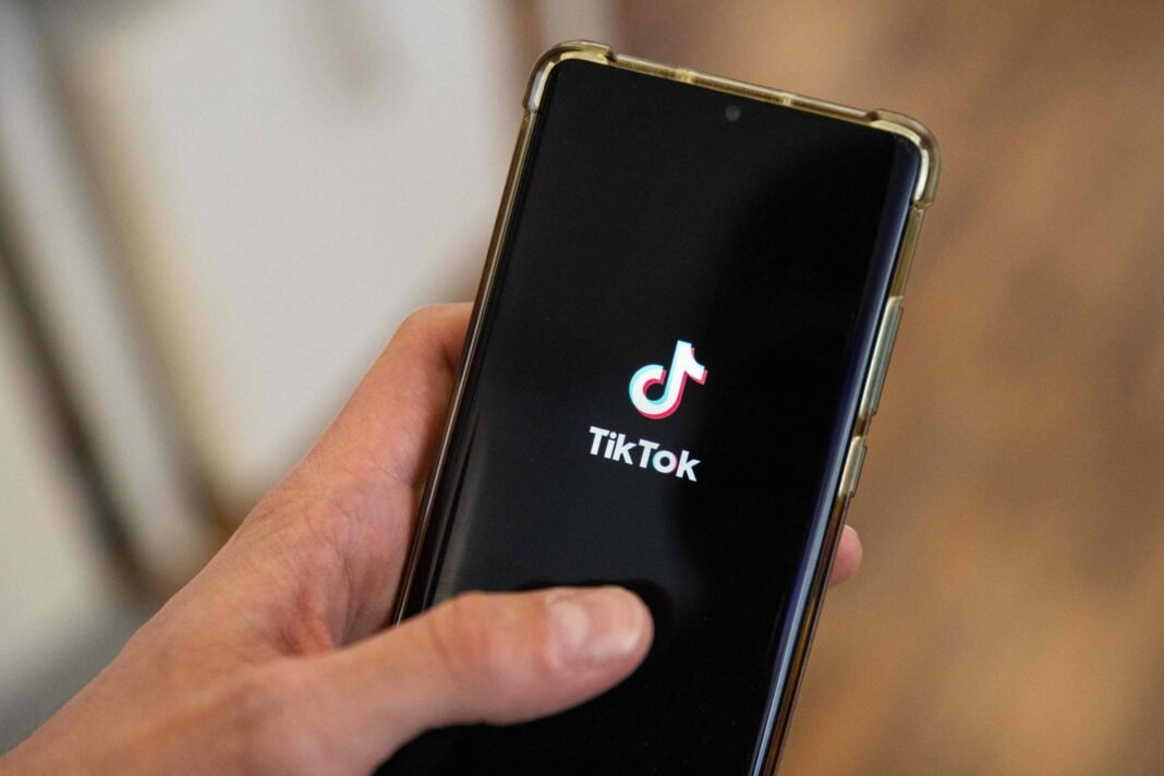 Mão segurando smartphone com tela exibindo o logo do TikTok em fundo preto. Fundo desfocado sugere ambiente interno com piso de madeira.