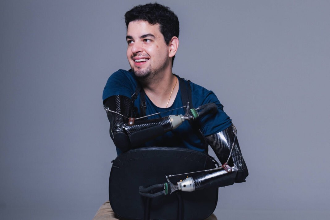 Homem jovem sentado de costas em cadeira preta, com braços e pernas substituídos por próteses robóticas pretas. Ele veste camiseta azul e calça bege, sorrindo e olhando para o lado.