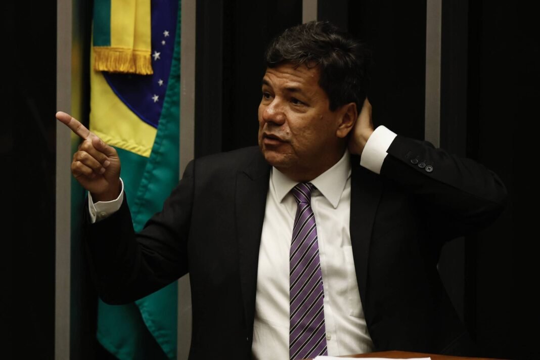 Homem de terno preto e gravata lilás gesticula com a mão direita apontando para o lado esquerdo, enquanto a mão esquerda toca a nuca. Ao fundo, bandeira do Brasil parcialmente visível.