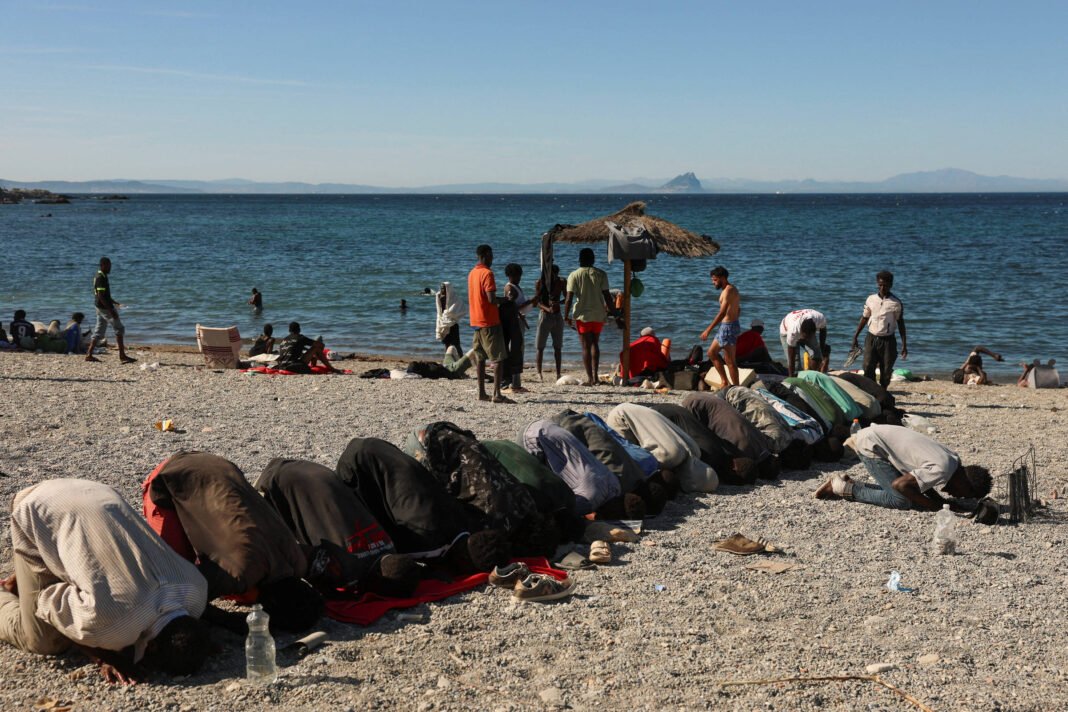 Dez pessoas ajoelhadas em fila na areia da praia, com a cabeça tocando o chão em posição de oração. Ao fundo, outras pessoas caminham e estão próximas à água do mar sob céu claro.