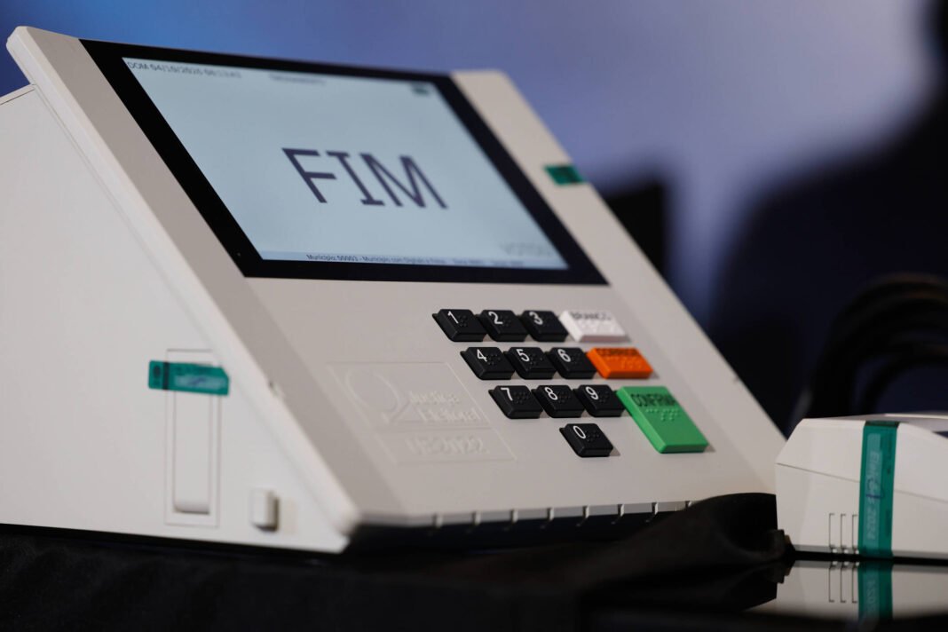 Urna eletrônica branca com teclado numérico e botões coloridos, exibindo a palavra 