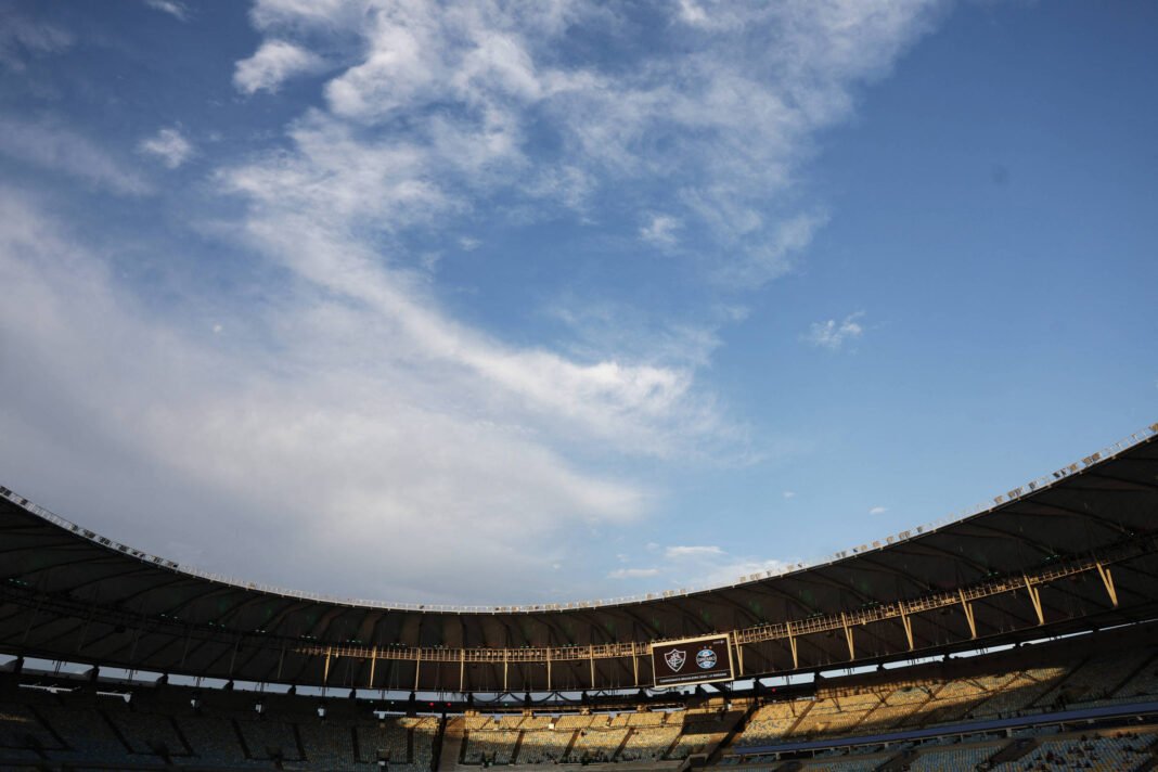 Vista ampla do interior de um estádio de futebol vazio com arquibancadas e cobertura parcial. O céu azul com nuvens brancas ocupa a maior parte da imagem acima da estrutura.