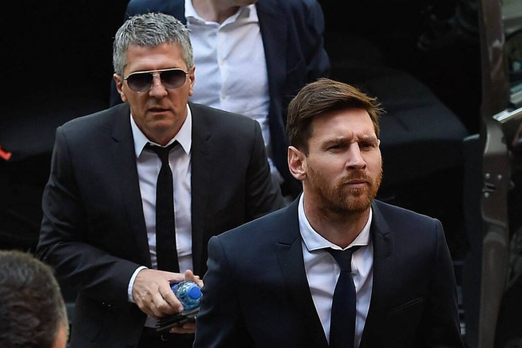 Dois homens vestidos com ternos escuros e gravatas pretas caminham em ambiente externo. O homem à frente tem barba e cabelo curto, enquanto o homem atrás usa óculos escuros e segura uma garrafa de água.