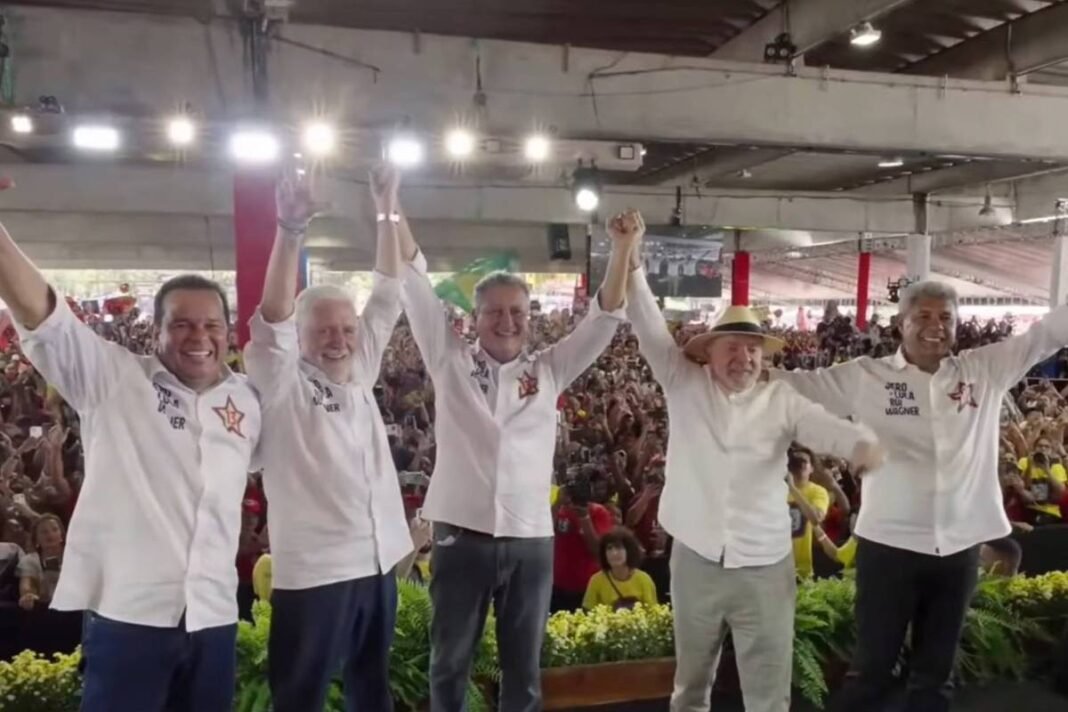 Cinco homens de camisa branca levantam as mãos unidas em sinal de vitória em um palco coberto. Atrás deles, multidão numerosa com camisetas vermelhas e amarelas acompanha o evento em ambiente interno com iluminação artificial.