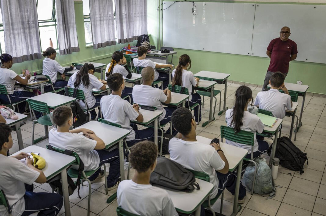 Vinte estudantes sentados em carteiras organizadas em fileiras dentro de sala de aula. Todos usam uniforme branco e azul, alguns com mochilas ao lado. Um professor está em pé próximo ao quadro branco na frente da sala, falando para a turma. A sala tem paredes verdes claras, janelas com cortinas e piso de cerâmica.