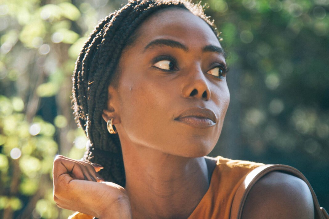 Mulher jovem de pele escura com tranças presas, usando blusa laranja sem mangas, olha para o lado direito. Fundo desfocado com vegetação e luz natural.