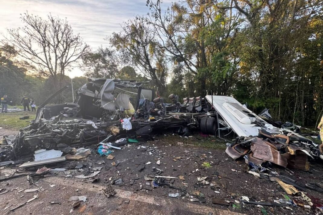 Caminhão tombado e destruído em estrada cercada por árvores. Detritos espalhados pelo chão e pessoas ao fundo observam a cena.