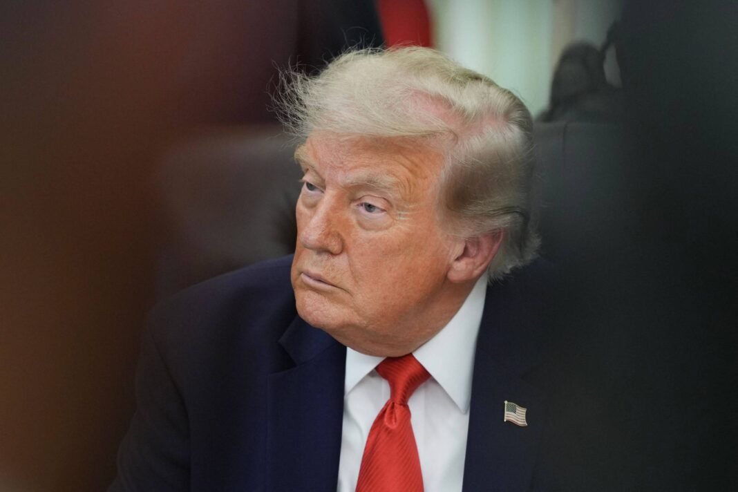 Homem com cabelo loiro claro penteado para trás, veste terno escuro, camisa branca e gravata vermelha, olhando para o lado direito em ambiente interno com fundo desfocado.