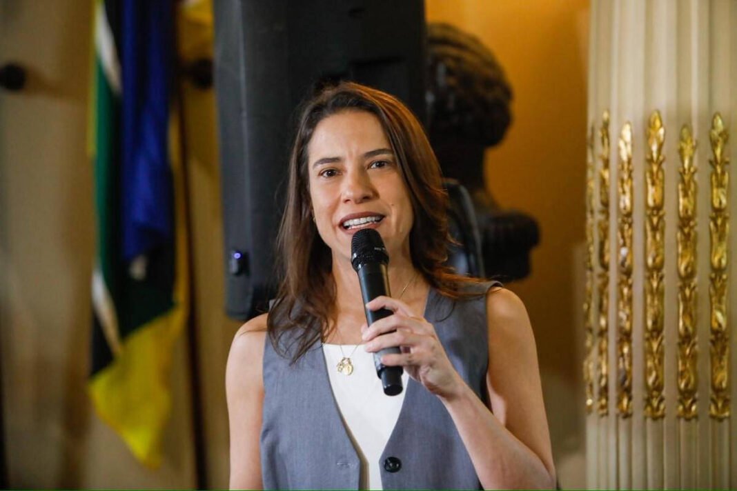 A governadora Raquel Lyra, mulher de cabelos castanhos e lisos segura microfone com a mão direita enquanto fala. Ela veste colete cinza sobre blusa branca. Ao fundo, bandeira com cores verde, azul e amarelo e detalhes dourados em coluna decorativa.
