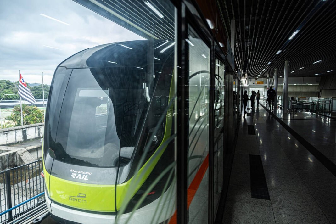 Trem do Aeroporto de Guarulhos parado na plataforma da estação, com reflexo no vidro da plataforma. Pessoas caminham no corredor interno da estação, e a bandeira do estado de São Paulo é visível ao fundo.