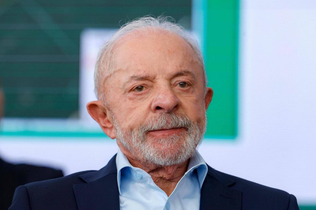 Homem idoso com cabelo branco e barba grisalha veste camisa azul clara e paletó escuro, sentado em ambiente interno com fundo desfocado.