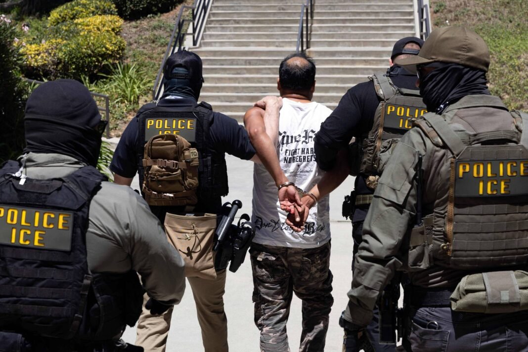 Quatro agentes da polícia ICE, com coletes táticos e roupas escuras, escoltam homem de camiseta branca e calça camuflada com as mãos algemadas para trás, subindo escadaria externa em área com vegetação.