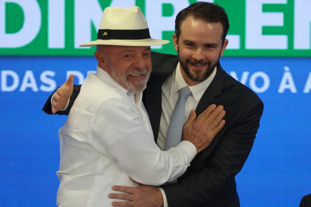 Homem mais velho de camisa branca e chapéu branco abraça homem mais jovem de terno preto e gravata cinza. Fundo azul com texto parcialmente visível.