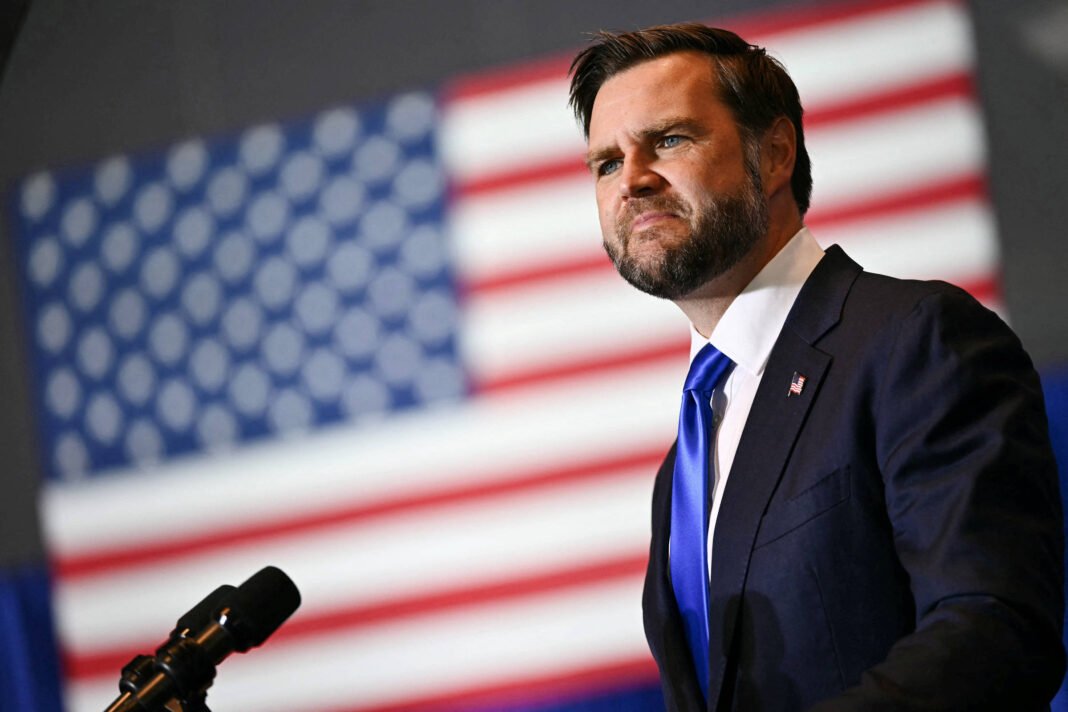 Homem de terno azul escuro, camisa branca e gravata azul fala em microfone com bandeira dos Estados Unidos desfocada ao fundo.