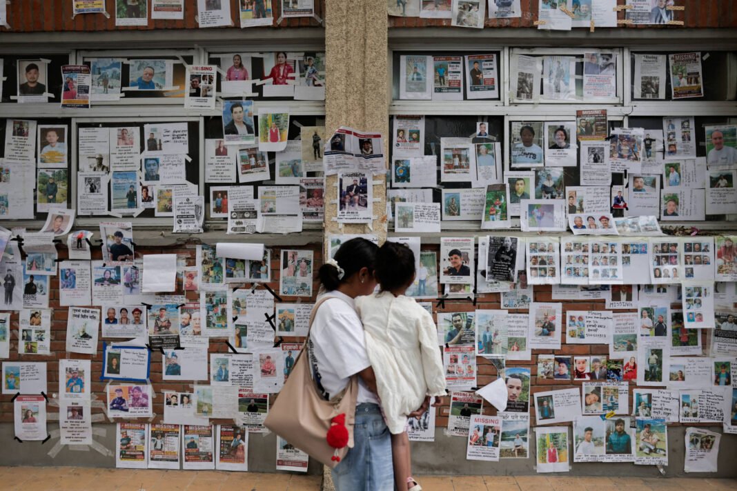Mural externo coberto por dezenas de cartazes com fotos e informações de pessoas desaparecidas. Em primeiro plano, mulher carrega criança no colo, ambas de perfil, caminhando em frente ao mural.