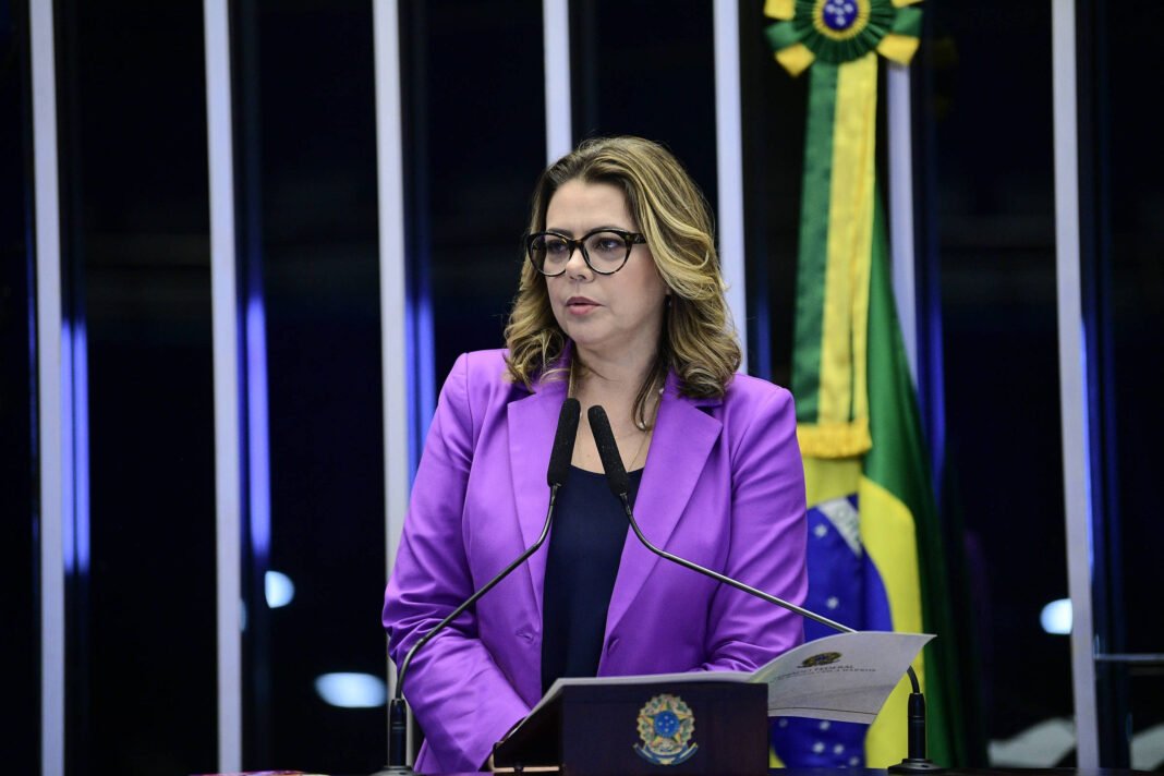 Senadora de cabelos claros e óculos fala em microfones no plenário. Ela veste blazer roxo e segura documento. Bandeira do Brasil está ao fundo.