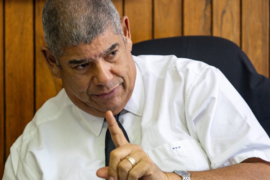 Um homem de pele clara, com cabelo curto e grisalho, está sentado em uma cadeira de escritório. Ele usa uma camisa branca e uma gravata escura. O homem está gesticulando com um dedo levantado, como se estivesse fazendo um ponto importante. Ao fundo, há uma parede de madeira com painéis. O ambiente parece ser um escritório.