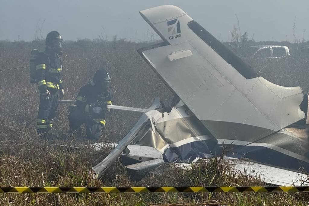 Destroços de avião pequeno com cauda visível em campo aberto. Dois bombeiros com equipamentos de proteção estão próximos, avaliando a cena. Fita amarela de isolamento delimita a área. Céu nublado ao fundo.