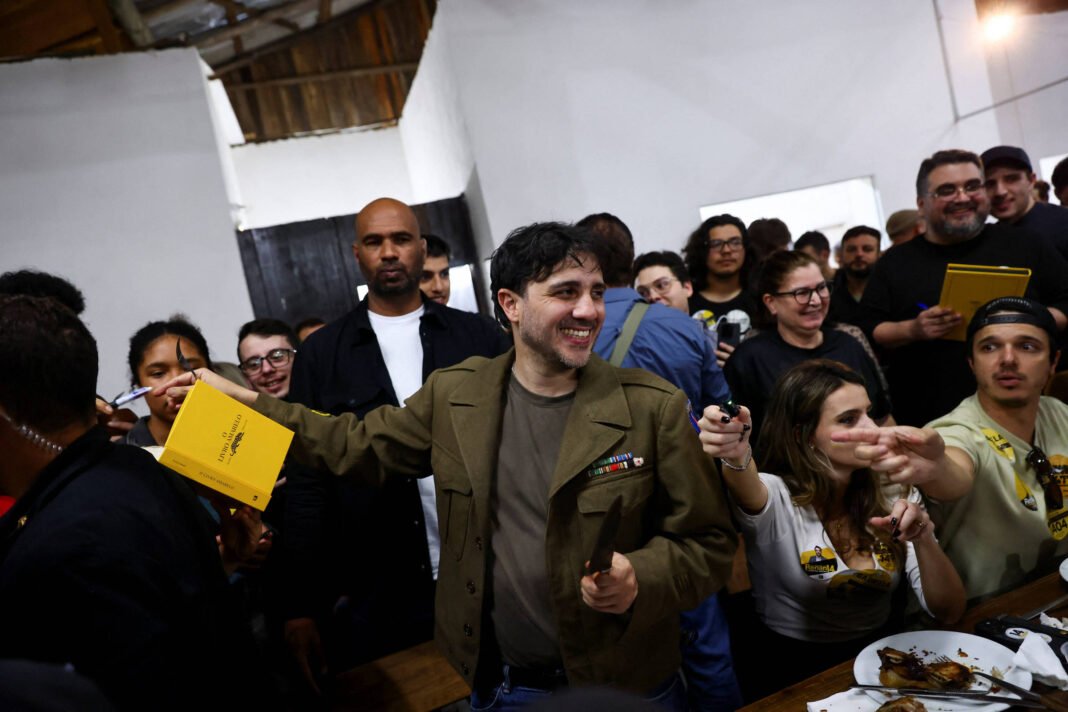 Homem sorridente com jaqueta verde distribui autógrafos em caderno amarelo para pessoas em ambiente interno com paredes brancas. Público ao redor observa e interage, alguns seguram canetas e celulares.