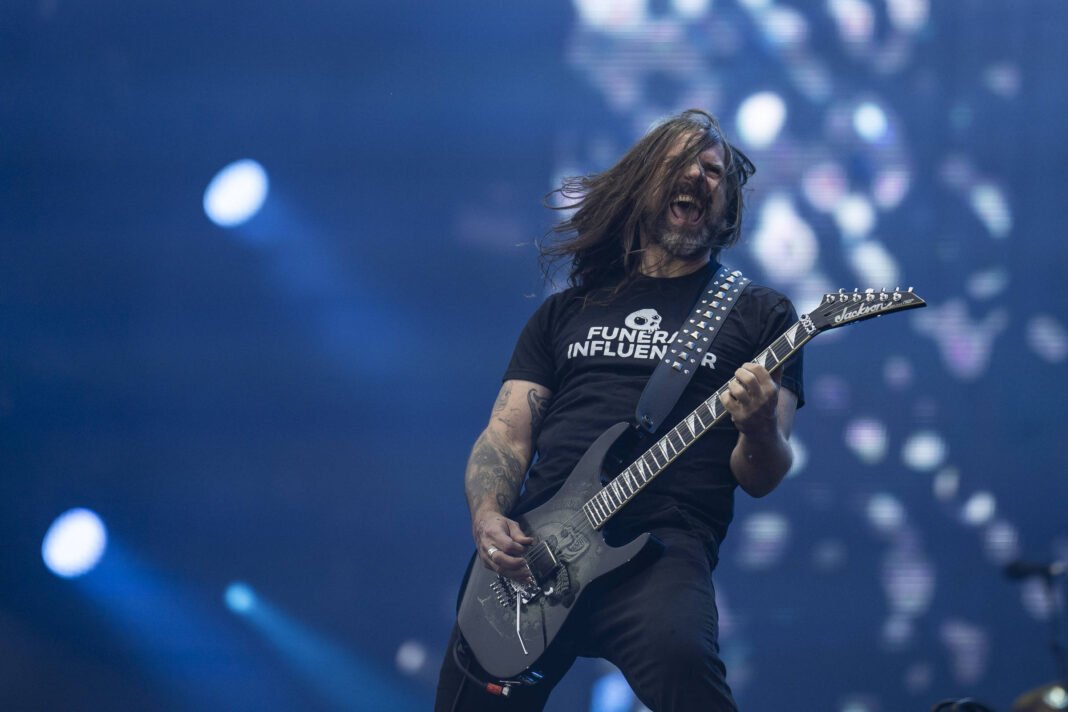 Guitarrista de cabelos longos toca guitarra elétrica em palco com iluminação azul e fundo desfocado. Ele veste camiseta preta com texto branco e está em postura enérgica.