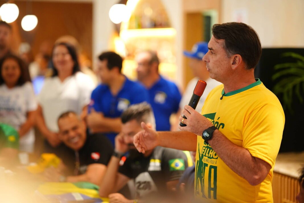 Homem de camiseta amarela com detalhes verdes segura microfone laranja e fala para grupo de pessoas em ambiente interno. Pessoas ao fundo observam e algumas sorriem, com iluminação quente e decoração simples.