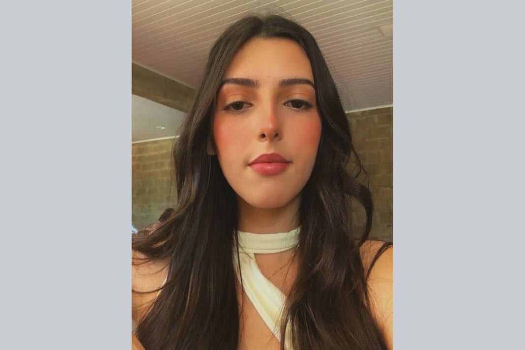 Mulher jovem com cabelo longo e escuro, usando blusa branca com detalhe no pescoço, posando em ambiente interno com parede de tijolos ao fundo.