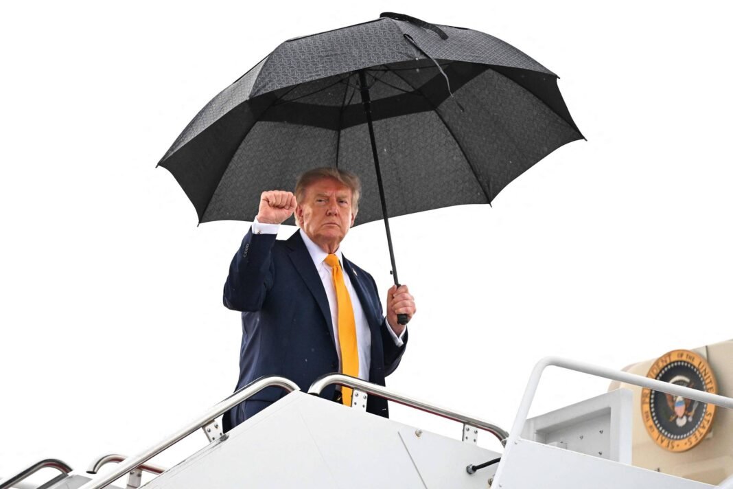 Homem de terno azul e gravata amarela segura guarda-chuva preto aberto enquanto está na escada de embarque de avião com selo presidencial visível.