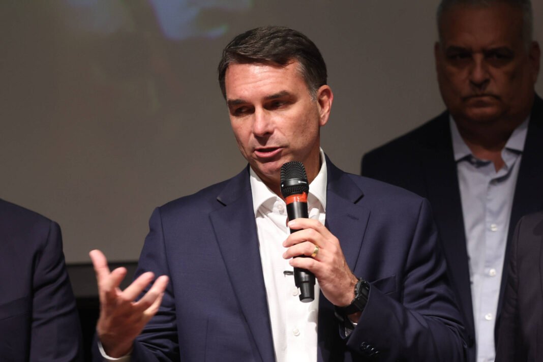 Quatro homens vestidos com ternos escuros e camisas claras estão alinhados em palco. O segundo da esquerda para a direita segura um microfone e fala, gesticulando com a mão esquerda. Os demais observam atentamente. Fundo escuro sem elementos visíveis.