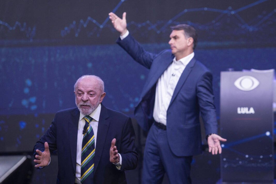 Dois homens em terno participam de debate em estúdio. À esquerda, homem mais velho com barba e gravata listrada fala gesticulando com as mãos. À direita, homem mais jovem em terno azul levanta os braços, com cadeira ao fundo marcada com o nome 