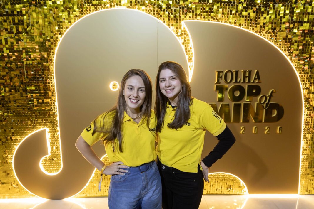 Duas mulheres posam sorrindo em frente a painel dourado iluminado com o logo do evento Folha Top of Mind 2020. Ambas vestem camisetas amarelas e estão lado a lado, com uma mão na cintura.