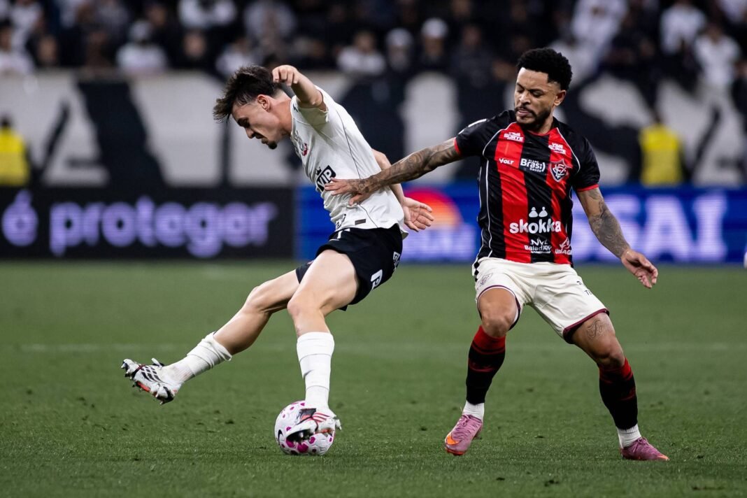 Dois jogadores disputam a posse da bola em campo durante partida noturna. Jogador de branco chuta a bola enquanto adversário de preto e vermelho tenta bloquear o lance.