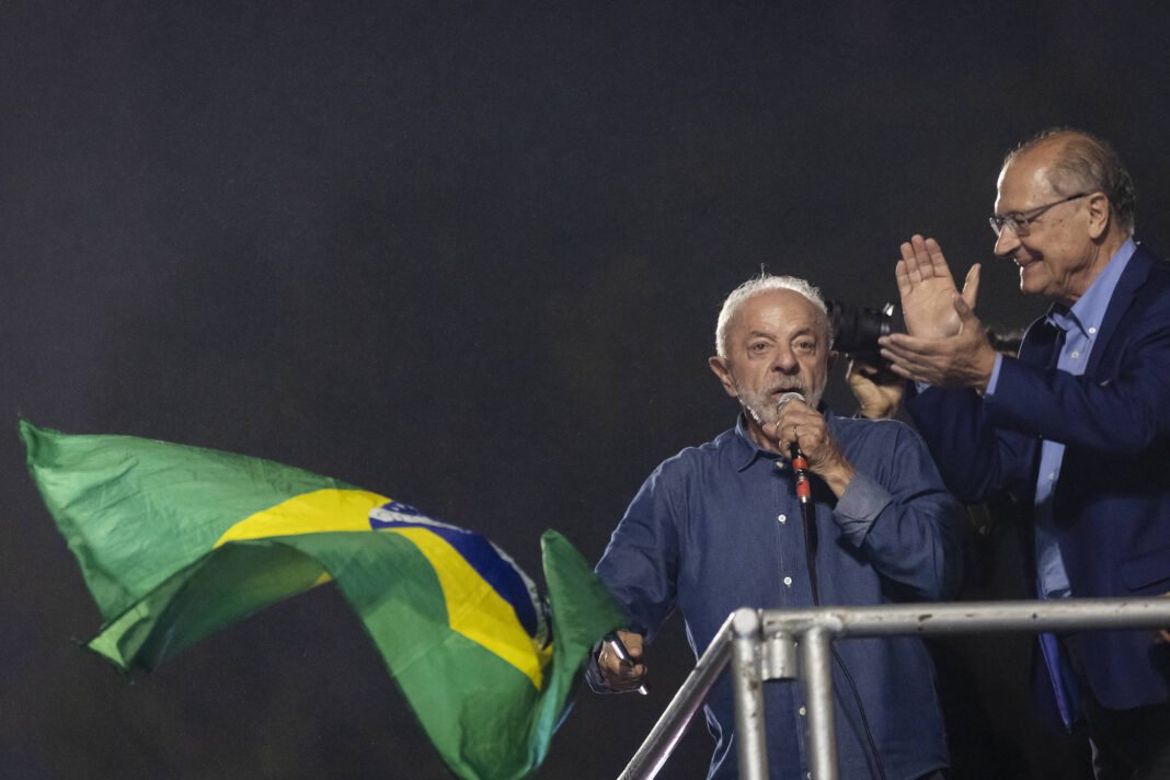 Homem de cabelos grisalhos segura e agita uma bandeira do Brasil enquanto fala ao microfone em um evento noturno. Outro homem ao lado aplaude, ambos em um palco com estrutura metálica.