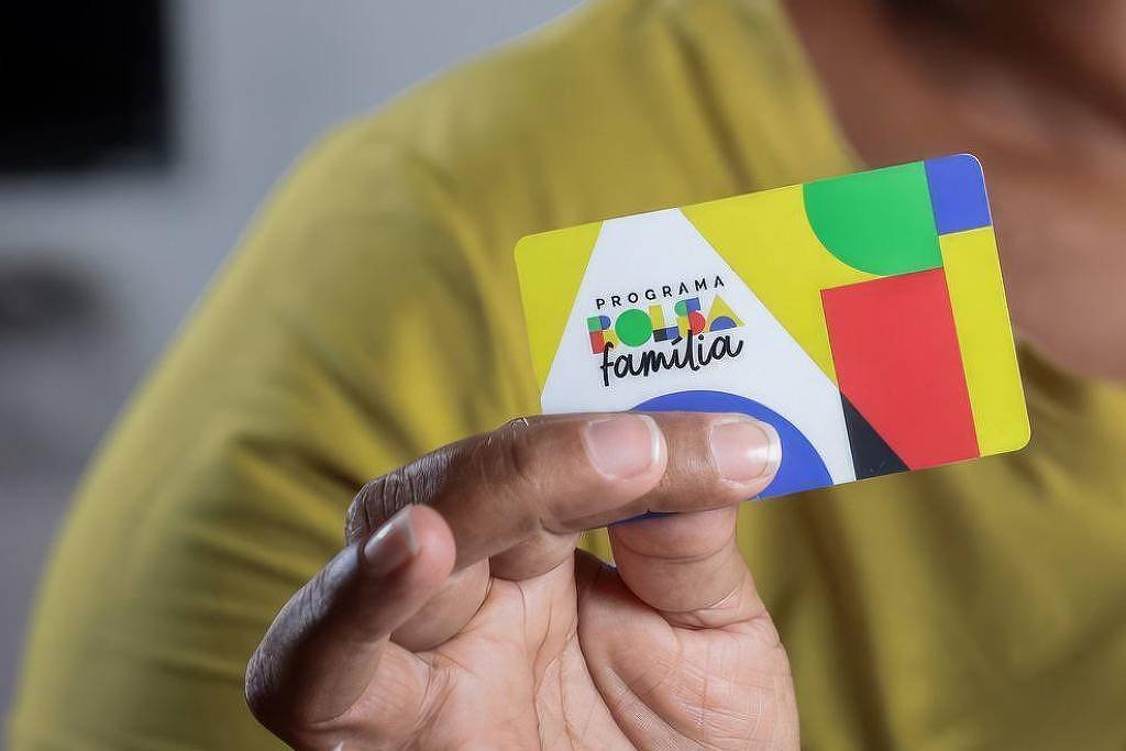 Mão de pessoa com camisa amarela segurando cartão colorido do Programa Bolsa Família, com design em verde, amarelo, azul e vermelho.
