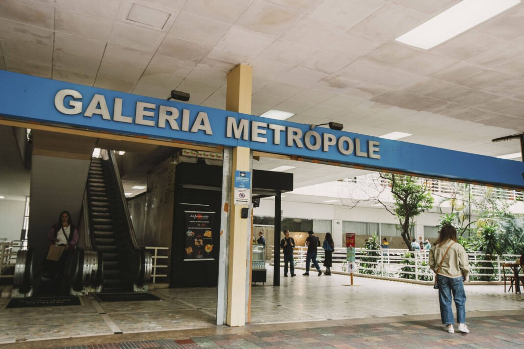 Entrada da Galeria Metrópole com placa azul e letras brancas. Escada rolante à esquerda com uma pessoa descendo. Pessoas caminham e estão paradas na área externa coberta, com árvores e bancos visíveis.