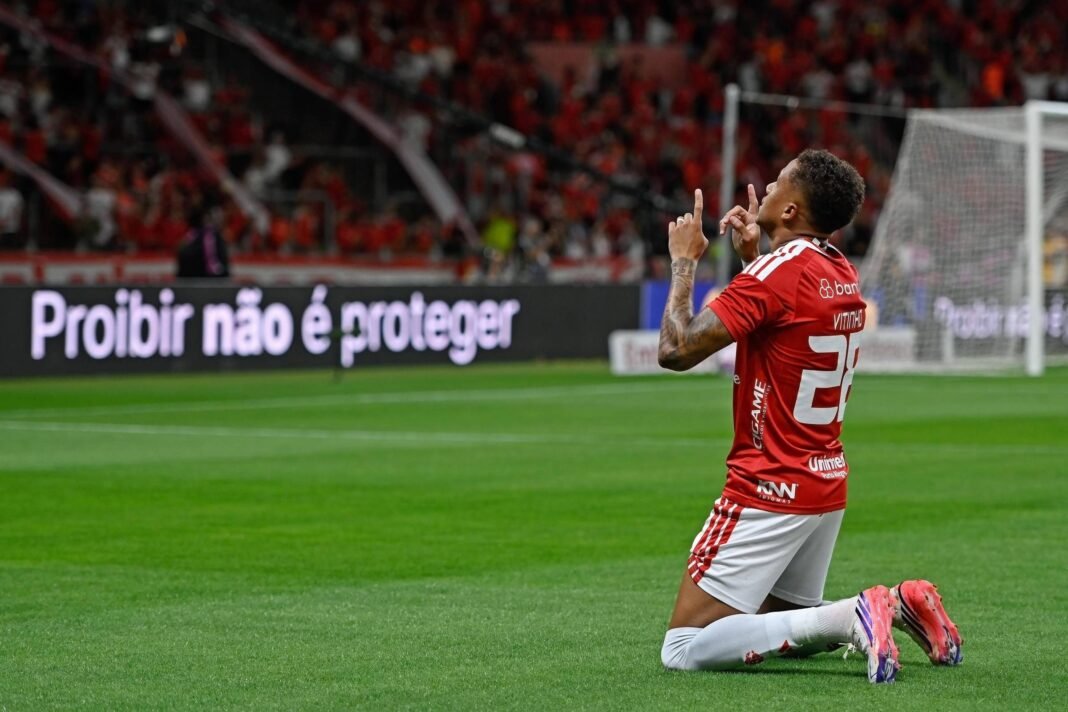 Jogador do Internacional ajoelhado no gramado do estádio Beira-Rio, com as mãos apontando para o céu em comemoração. Ao fundo, torcida nas arquibancadas e placa eletrônica com a frase 