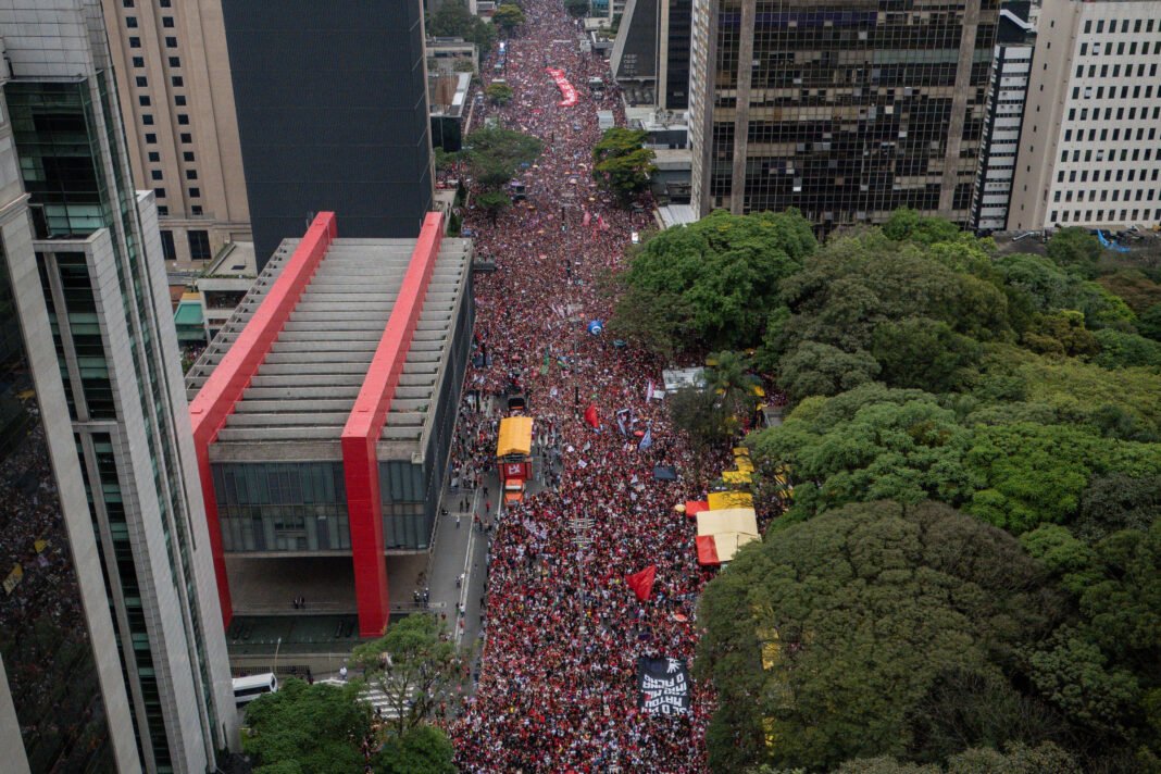 Multidão densa ocupa a Avenida Paulista entre prédios altos e áreas verdes em dia nublado. Pessoas vestem vermelho e seguram cartazes e bandeiras ao longo da via.