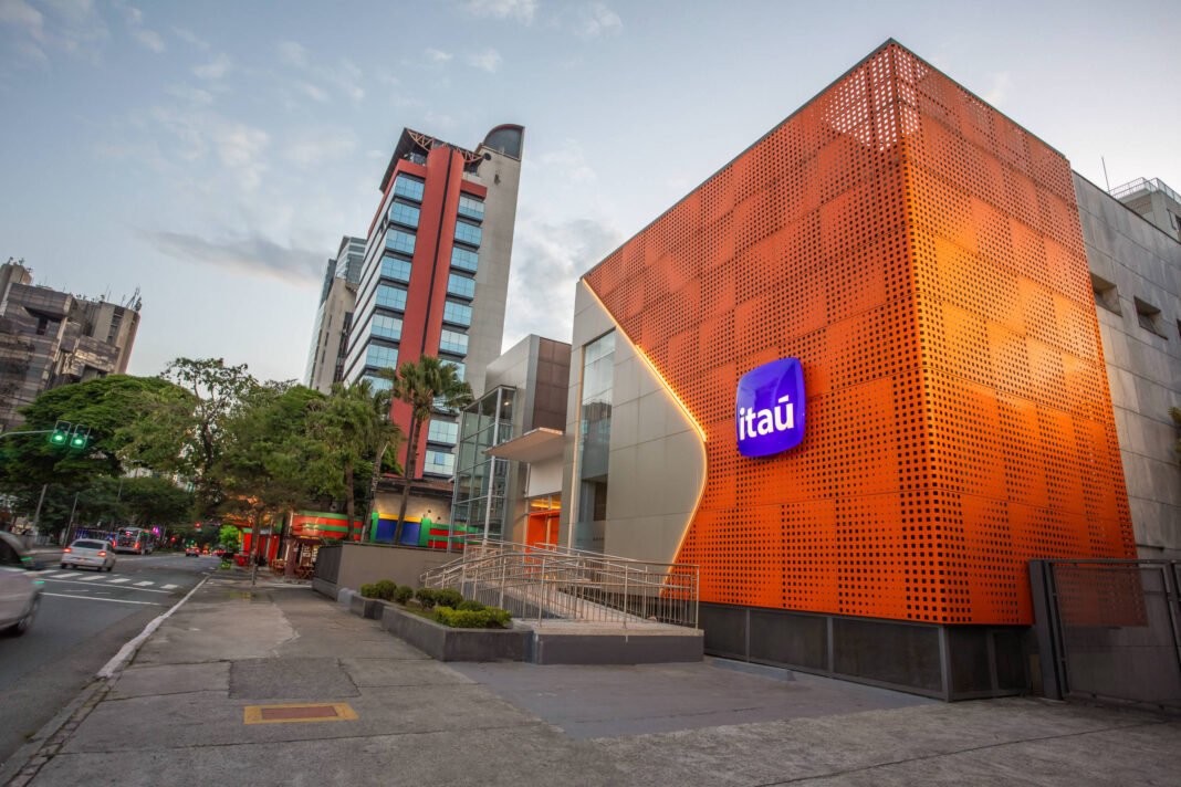 Fachada laranja iluminada de agência do banco Itaú em rua movimentada ao entardecer. Prédios altos e árvores cercam o local.