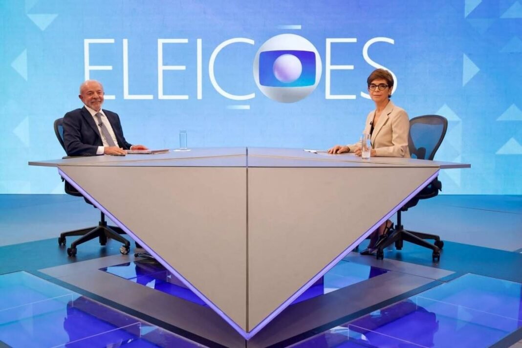 Dois apresentadores sentados em cadeiras giratórias em lados opostos de uma mesa triangular em estúdio com fundo azul e a palavra 