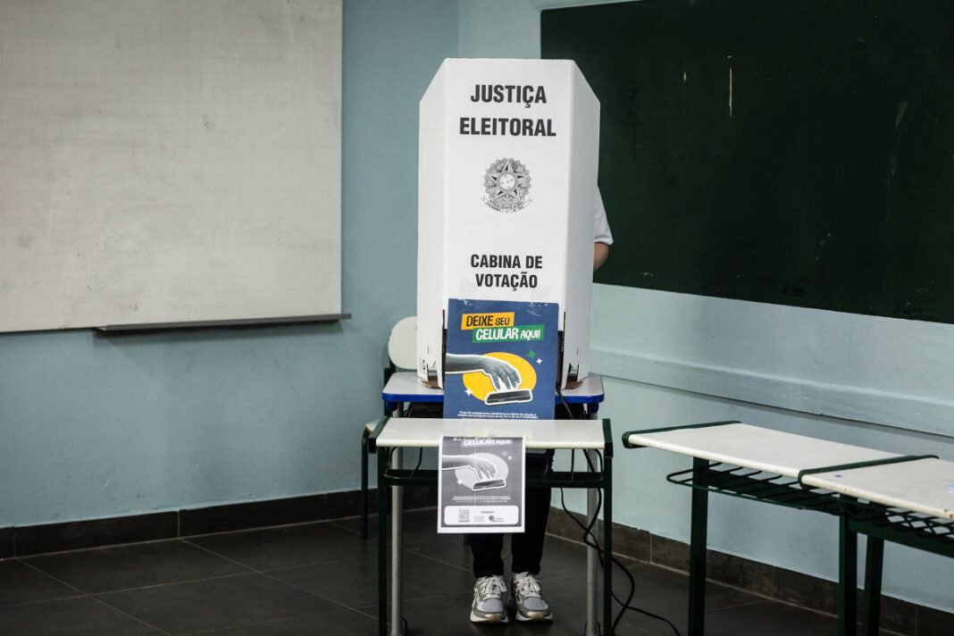 Pessoa em cabine de votação da Justiça Eleitoral dentro de sala de aula. Cabine branca com aviso para desligar celular, quadro negro ao fundo e carteiras escolares ao lado.