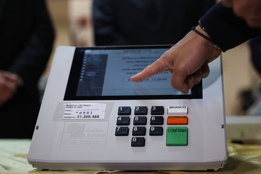 Mão aponta para a tela de urna eletrônica branca com teclado numérico e botões coloridos. Tela exibe interface de votação com texto e números. Fundo desfocado mostra pessoas em traje escuro.