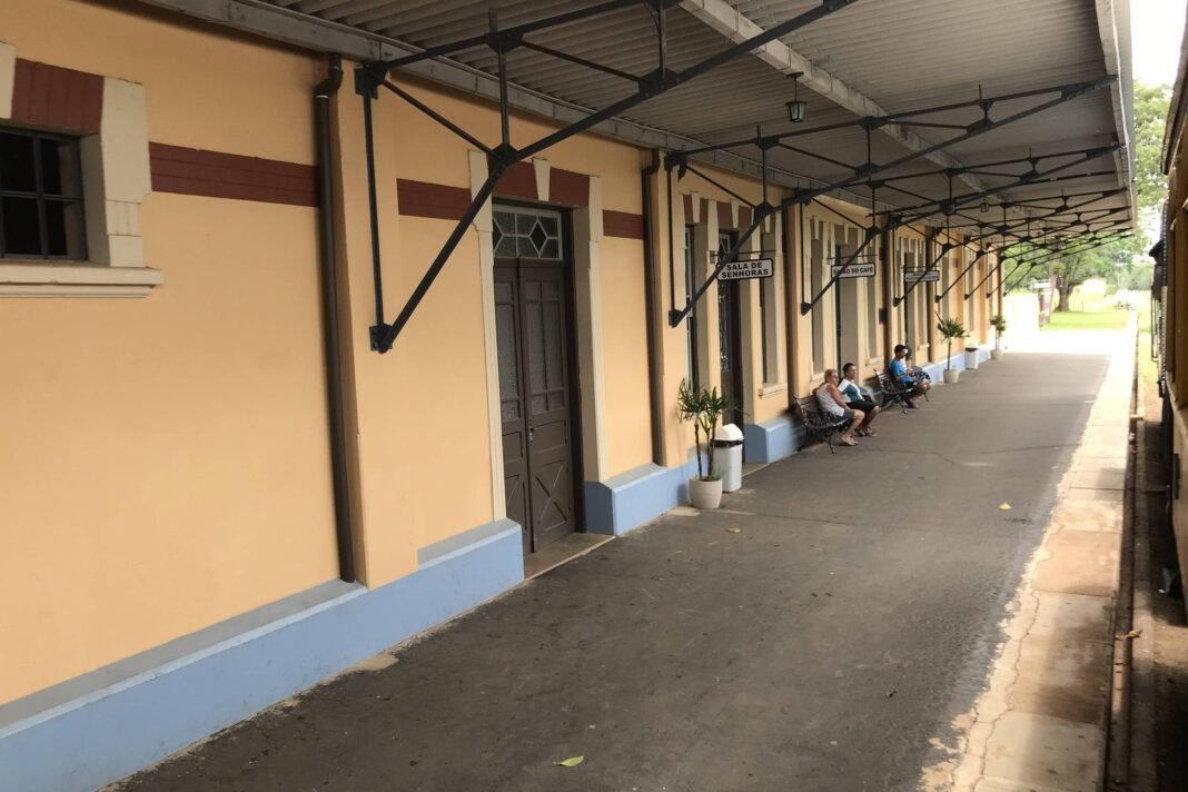 Plataforma longa de estação com cobertura metálica e paredes amarelas com detalhes marrons e azul claro. Cinco pessoas estão sentadas em bancos ao fundo, próximas a portas e janelas da estação.